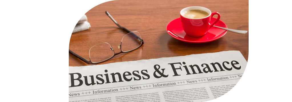 Auf dem Teakholztisch liegt eine Zeitung mit dem Titel "Business & Finance". Daneben eine Lesebrille und eine Tasse Espresso.