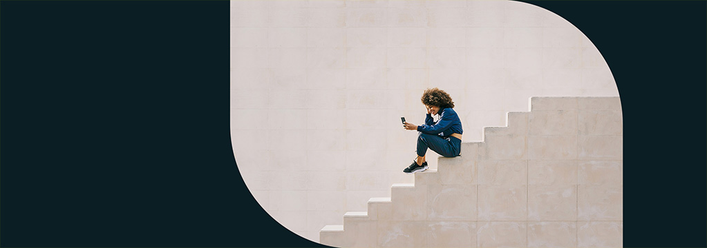 Eine Frau in blauer Trainingskleidung sitzt auf einer beigefarbenen Treppe und schaut erfreut auf ihr Smartphone.