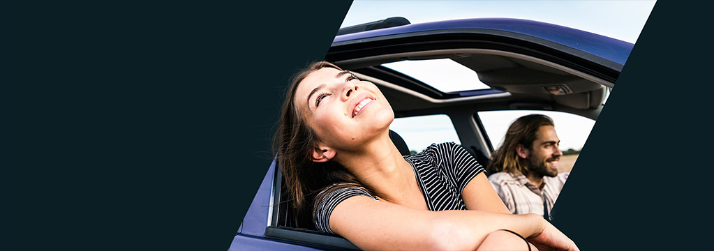Eine junge Frau lehnt sich l&auml;chelnd aus dem Fenster eines Autos, im Hintergrund ist der Fahrer zu sehen.