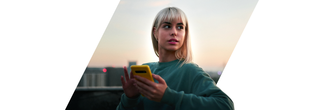 Eine junge Frau steht in der Abendsonne vor einer Skyline. Sie hält ihr gelbes Smartphone in der Hand und  möchte Festgeld anlegen.