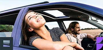Eine junge Frau lehnt sich l&auml;chelnd aus dem Fenster eines Autos, im Hintergrund ist der Fahrer zu sehen.
 