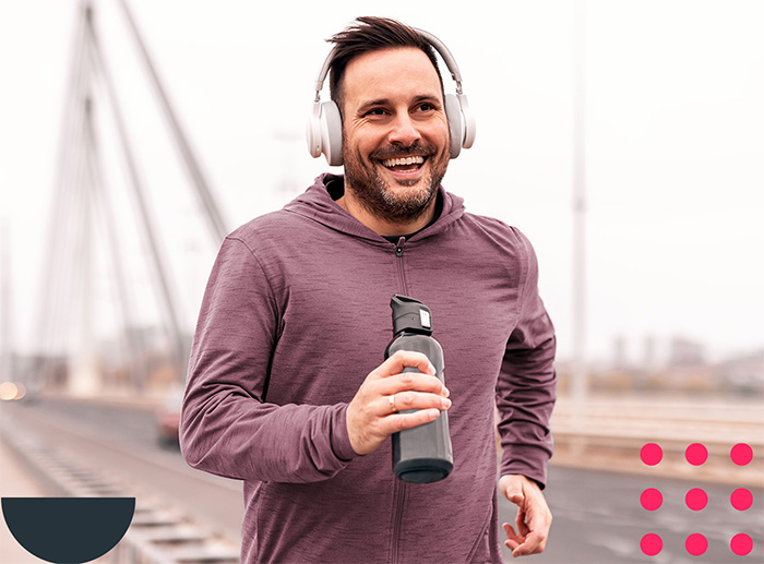 Joggender Mann mit Kopfh&ouml;rern und Trinkflasche l&auml;uft l&auml;chelnd &uuml;ber eine Br&uuml;cke.