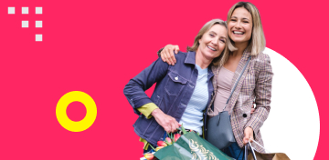 Zwei lachende Frauen mit vollen Einkaufstaschen umarmen sich.