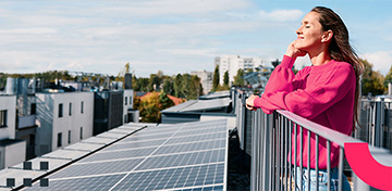Eine Frau steht entspannt auf einem Balkon neben Solarpaneelen in der Sonne.