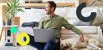Ein Mann mit Laptop sitzt vor einem Sofa und streichelt seinen Hund. Er freut sich über seine comdirect Depots.