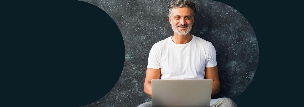 Ein Mann mit Bart sitzt mit seinem Laptop vor einer dunklen Wand.