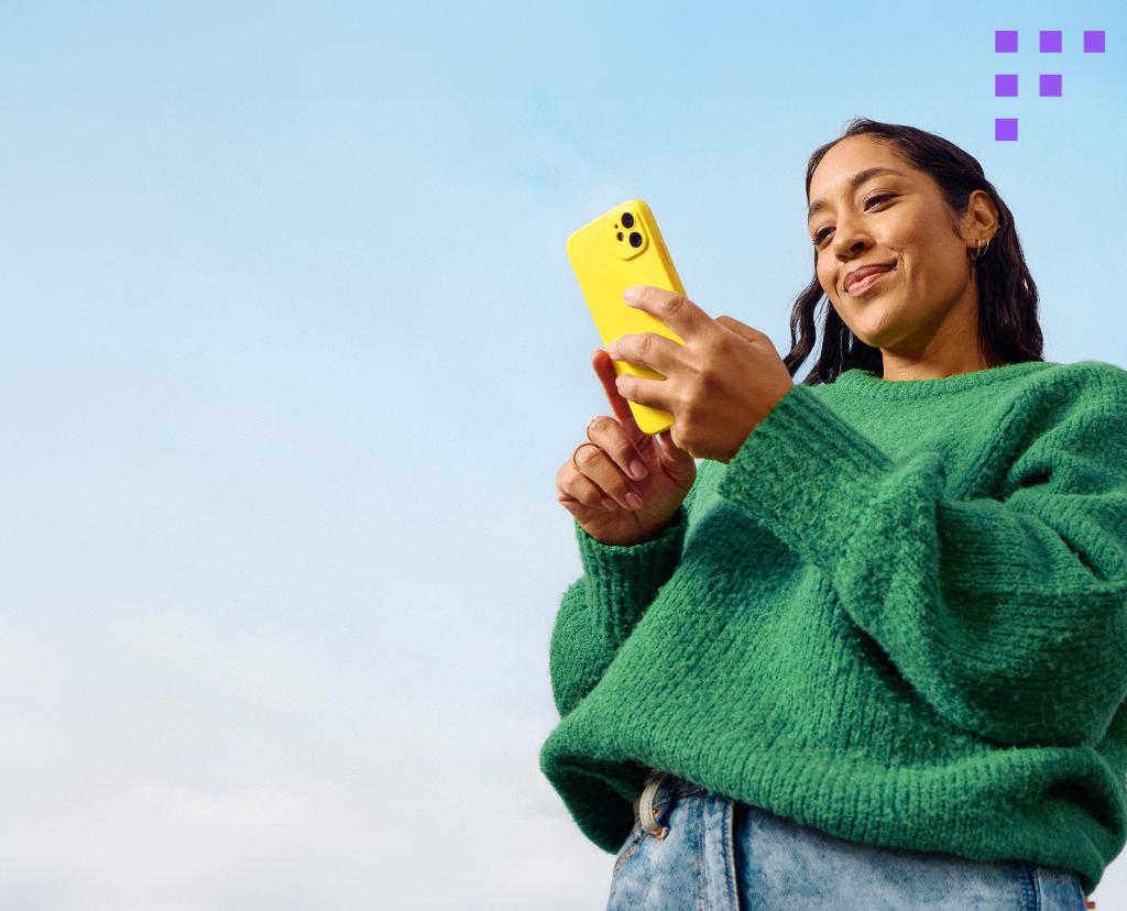 Eine junge Frau schaut gl&uuml;cklich auf ihr leuchtend-gelbes Smartphone.