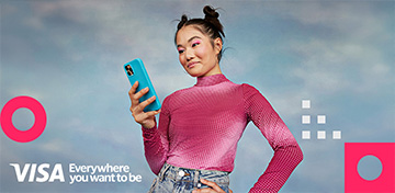 Eine junge Frau in rotem Oberteil h&auml;lt ein Smartphone und blickt auf das Display. Auf dem Bild ist das Logo von Visa mit dem Zusatz „Everywhere you want to be“ platziert.