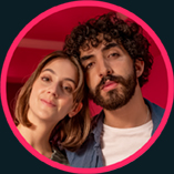 Zwei Personen stehen nebeneinander in ihrem Wohnzimmer. Eine Person trägt ein Hemd über einem T-Shirt.