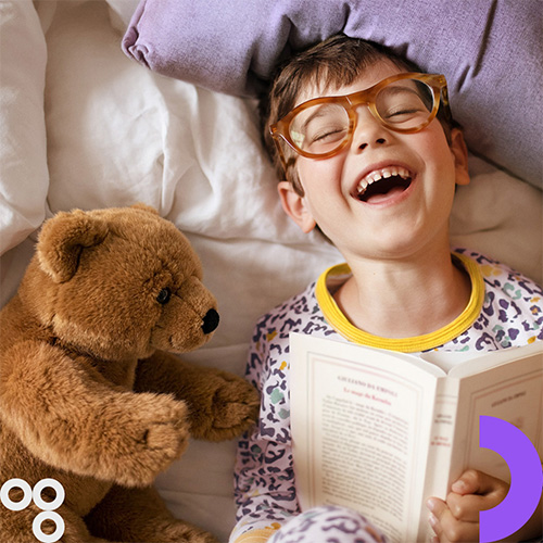 Ein lachender Junge mit Brille liest im Bett ein Buch. Neben ihm liegt ein Stofftier-B&auml;r. Es gibt den Hinweis „Bis 15.07.“.