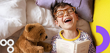 Ein lachender Junge mit Brille liest im Bett ein Buch. Neben ihm liegt ein Stofftier-B&auml;r. Es gibt den Hinweis „Bis 15.07.“.