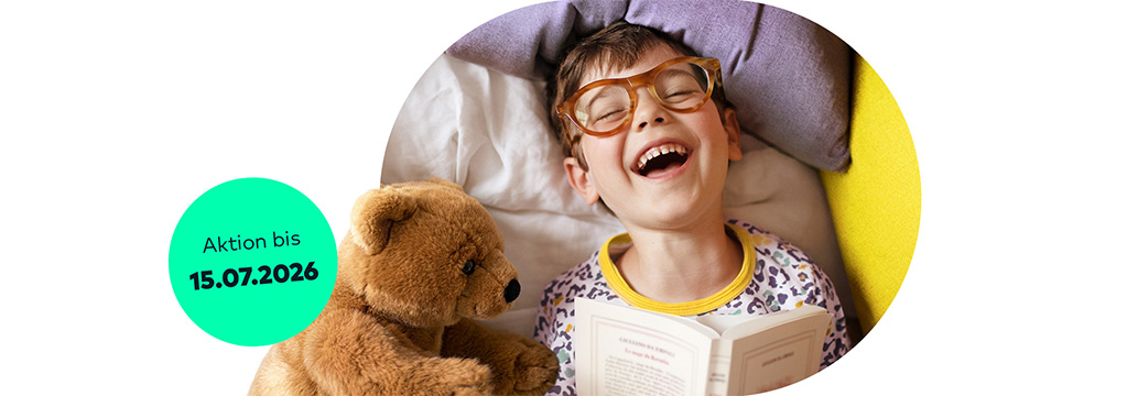 Ein lachender Junge mit Brille liest im Bett ein Buch. Neben ihm liegt ein Stofftier-B&auml;r. Auf einem St&ouml;rer steht „Aktion bis 15.07.2026“.