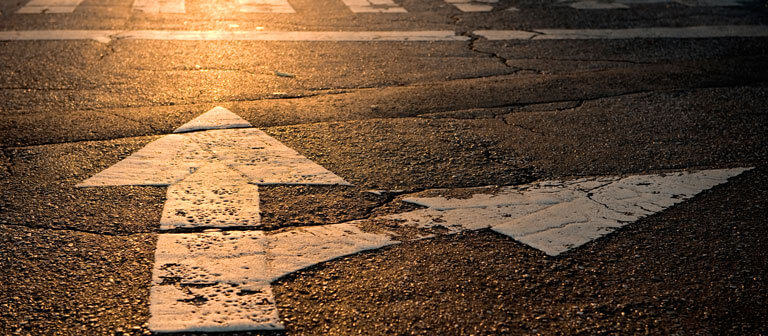 Auf einer laternenbeschienenen Straße sind weiße Richtungspfeile zu sehen.