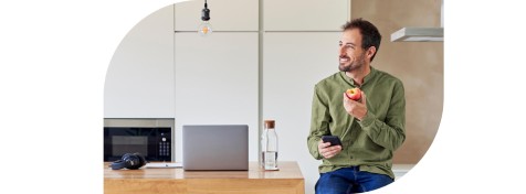 Ein Mann sitzt entspannt in seiner Küche. Er hält einen Apfel und ein Smartphone in den Händen.