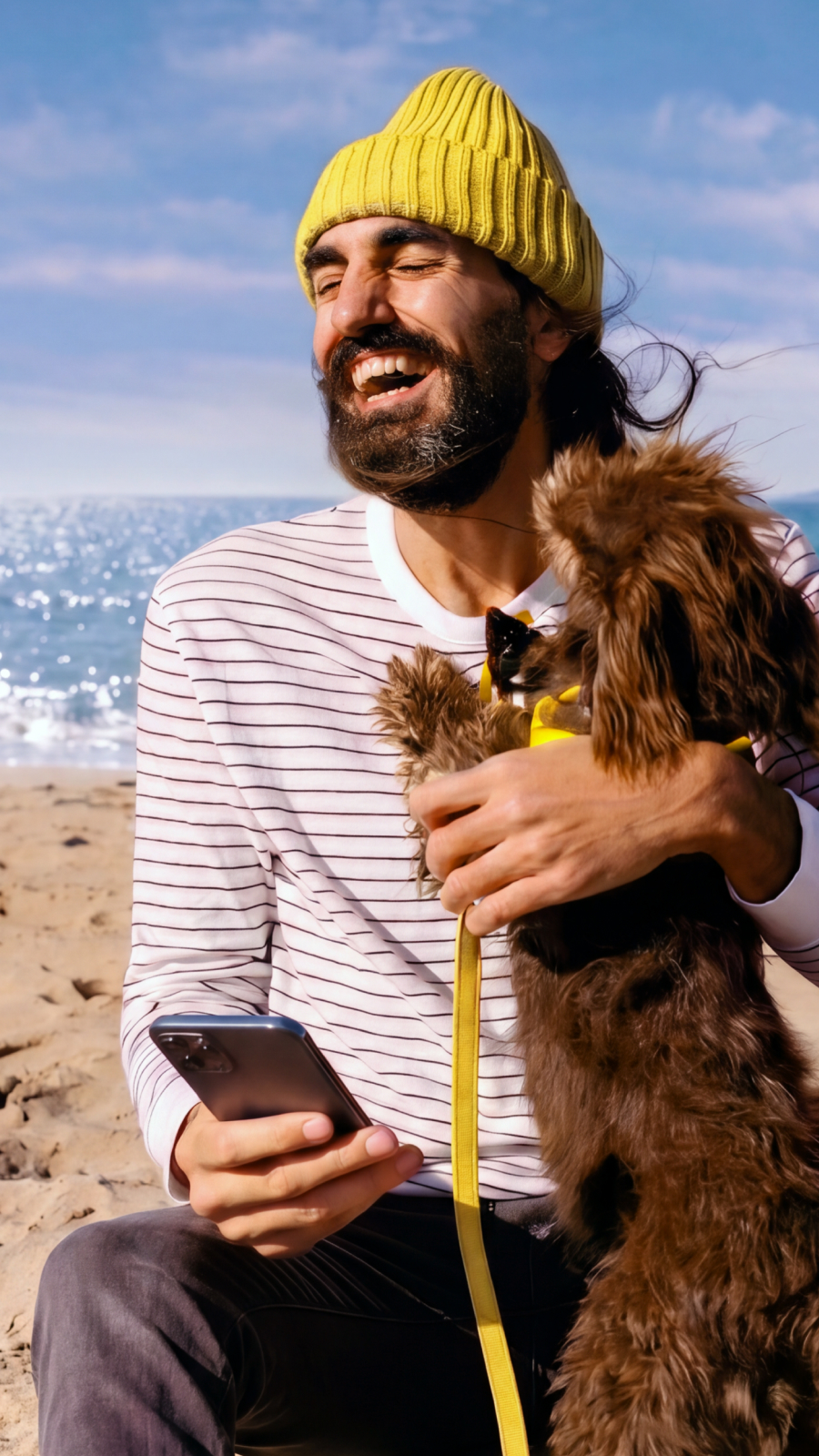 Ein lachender Mann mit gelber M&uuml;tze sitzt am Strand, h&auml;lt ein Smartphone in der Hand und umarmt einen kleinen Hund.