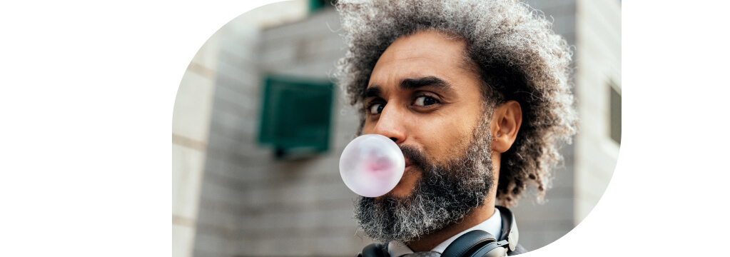 Ein lockiger Mann blickt verschmitzt in die Kamera und macht eine rosa Kaugummiblase.