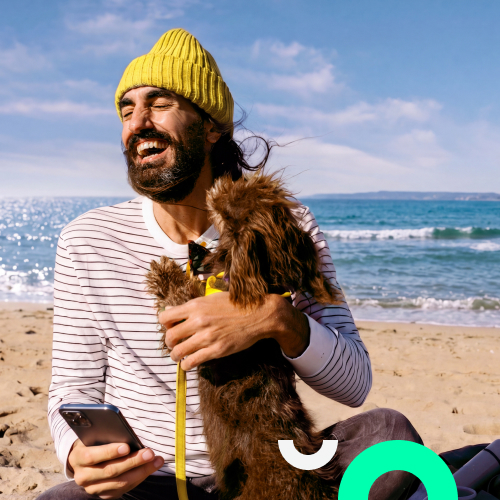 Ein lachender Mann mit gelber M&uuml;tze sitzt am Strand, h&auml;lt ein Smartphone in der Hand und umarmt einen kleinen Hund.