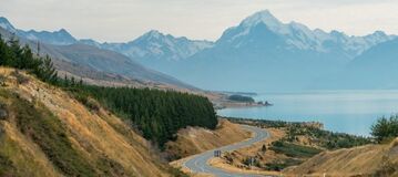 Eine asphaltierte Stra&szlig;e, die an einem ruhigen See vorbeif&auml;hrt, mit einer majest&auml;tischen Bergkette im Hintergrund.