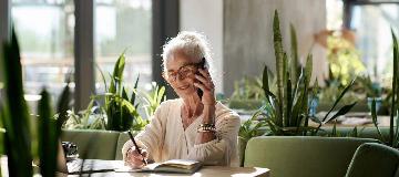 Eine Frau sitzt telefonierend in einem Café und macht sich Notizen.