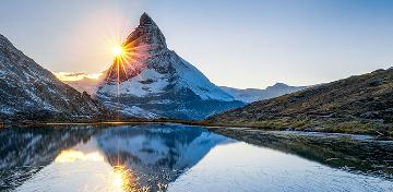 Die tiefstehende Sonne und ein schneebedeckter Berg spiegeln sich in einem See in den Schweizer Alpen.