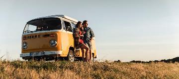 Ein junges Paar lehnt entspannt an einem gelben Transporter und schaut in die Ferne.