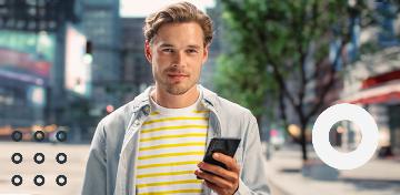 Ein junger Mann h&auml;lt ein Smartphone in der Hand l&auml;chelt freundlich.
