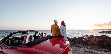 Ein junges Paar lehnt an einem roten Cabrio und schaut aufs Meer.