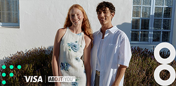 Eine modisch gekleidete Frau und ein modisch gekleideter Mann stehen in einem modernen Geschäft und freuen sich über 15 % Rabatt von Visa und About You.