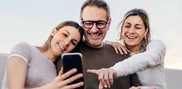 Vater, Mutter und Tochter sitzen l&auml;chelnd zusammen und schauen auf ein Handy, das die Tochter h&auml;lt.