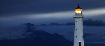 Ein weißer Leuchtturm hebt sich vor einem dunklen Himmel und einem wolkenverhangenen Berg ab.