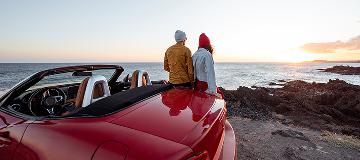 Ein junges Paar lehnt an einem roten Cabrio und schaut aufs Meer.