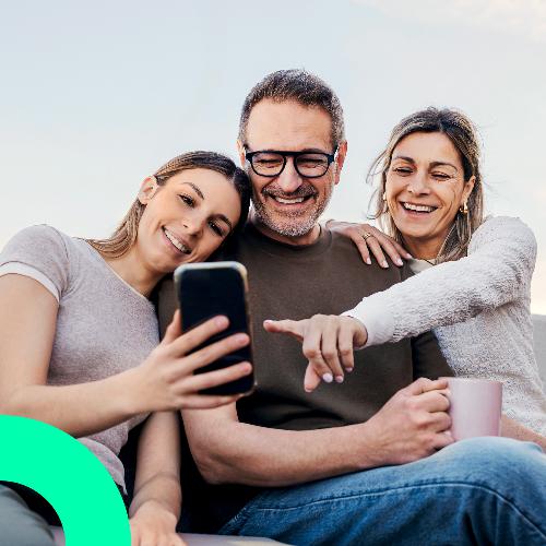 Eine Familie sitzt l&auml;chelnd zusammen und schaut gemeinsam auf ein Smartphone.